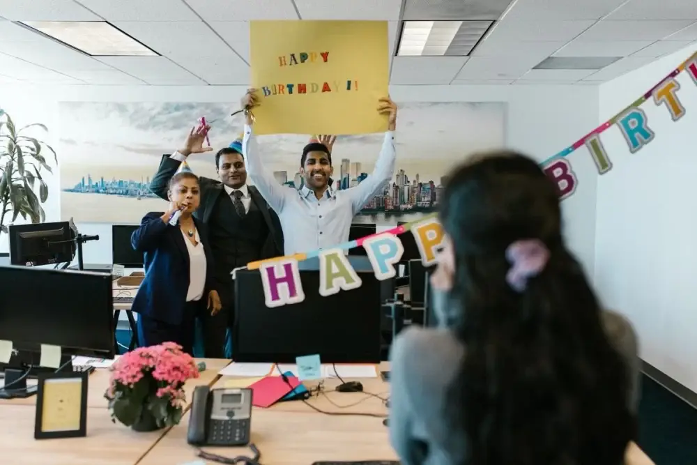 Three coworkers celebrate with a “Happy Birthday” sign in a Barcelona office—unique employee recognition by MIT Creators, expert DMC events.